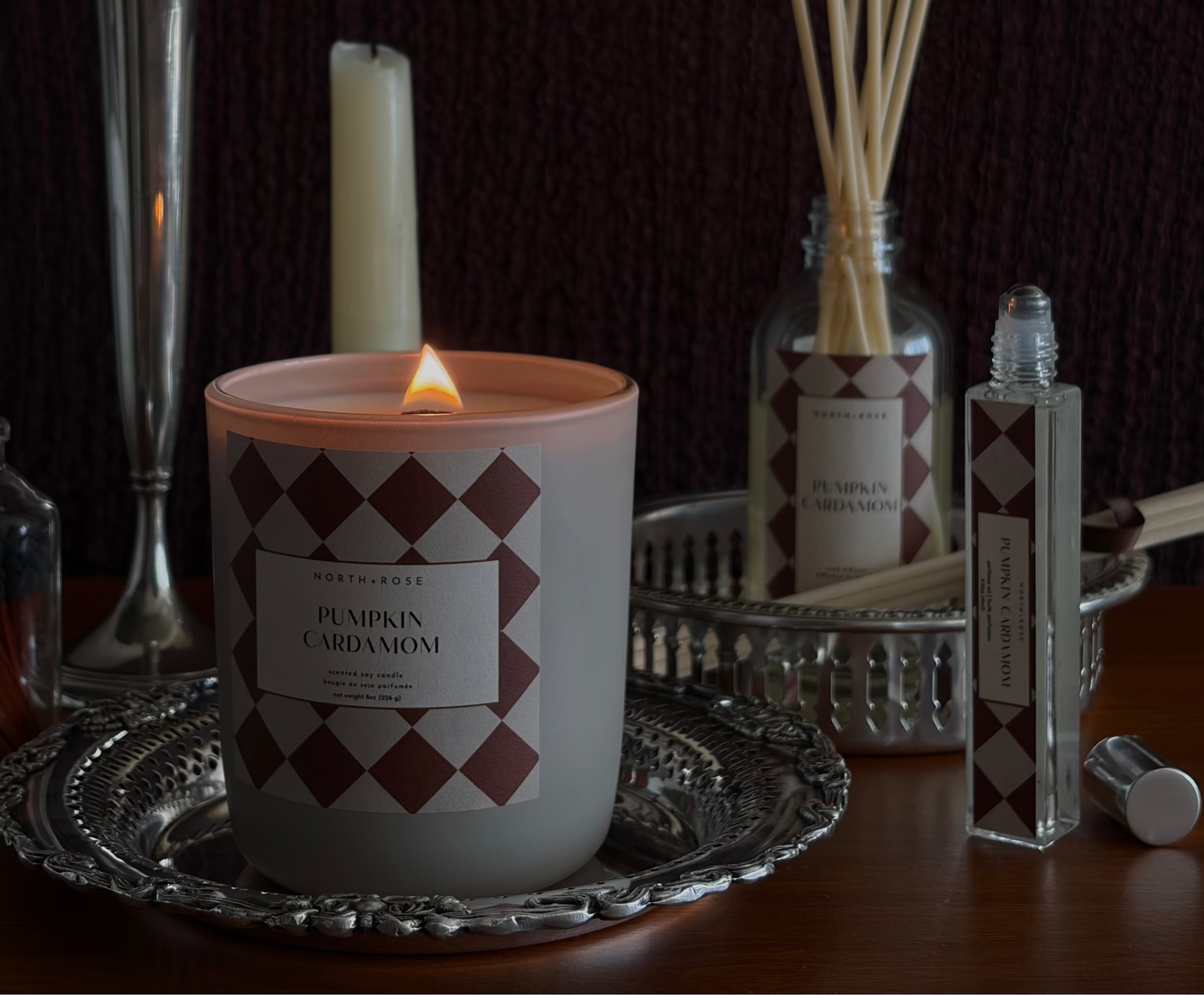 A lit candle in a white jar with a burgundy and white diamond pattern, styled on a silver tray alongside a reed diffuser, a diffuser bottle, a candlestick with a tall white candle, and a small bottle with its cap off, all set against a dark textured background.