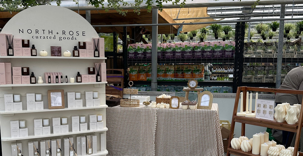 A styled product display inside a greenhouse or botanical nursery, featuring a large cream-colored arched shelf labeled 'NORTH-ROSE GOURMET' filled with boxed and bottled products, a central table with a beige checkered cloth, and a wooden two-tier shelf with white candles and ceramic items.